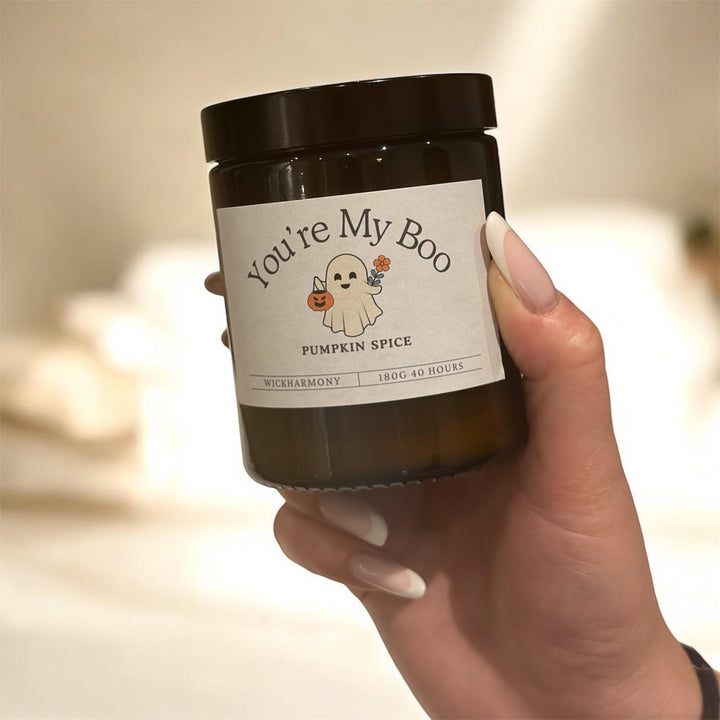 Hand holding a pumpkin spice candle labeled 'You're My Boo' with a ghost and pumpkin design.