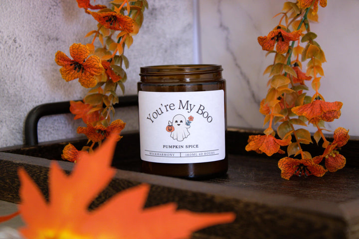 Candle labeled 'You're My Boo' with pumpkin spice scent on a surface with decorative flowers.