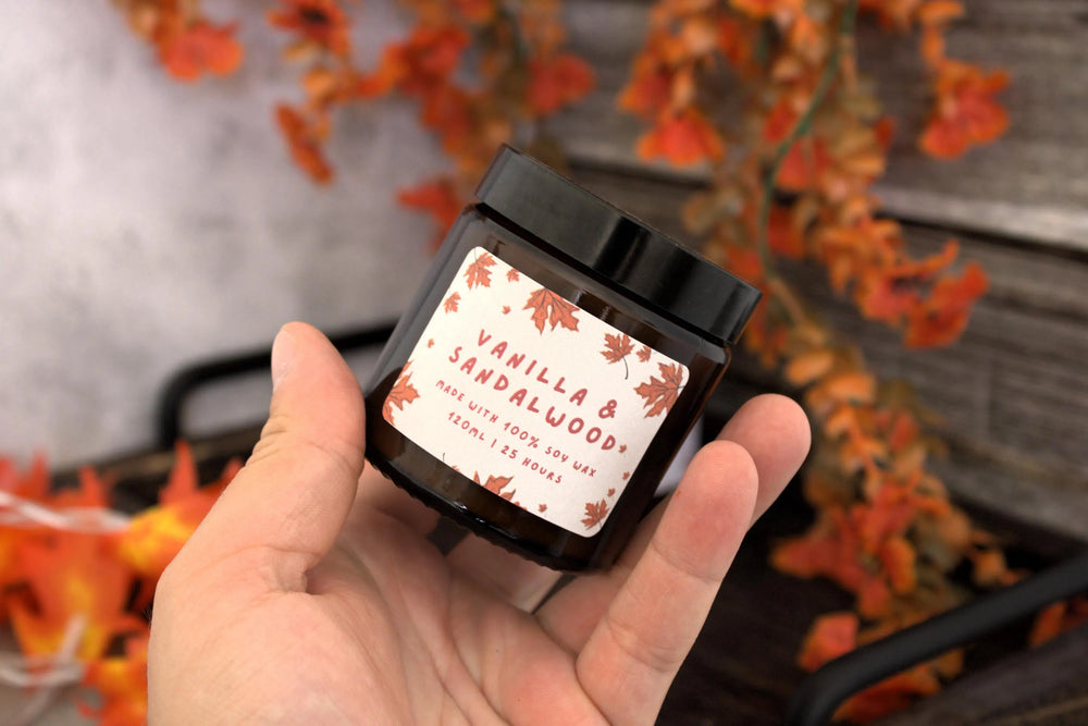 Hand holding a vanilla sandalwood air freshener with a blurred autumn leaf background